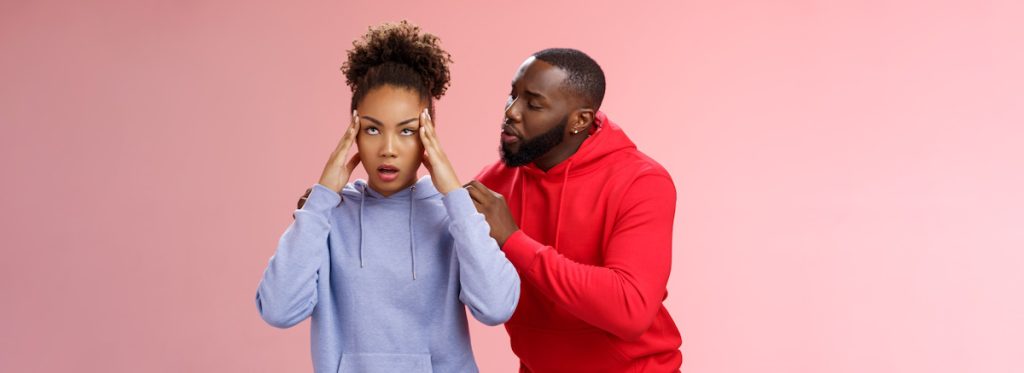 Man whispering to unimpressed woman on pink background.