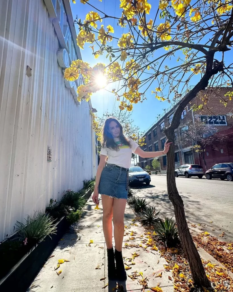 Jamie Clayton strolling under blooming tree on sunny street.